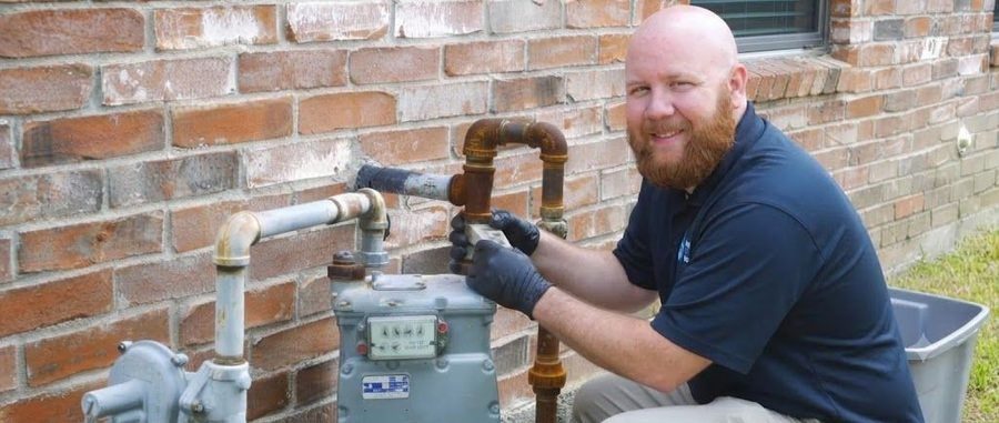 Bearded maintenance worker in a navy shirt and gloves kneels beside an outdoor gas meter, turning the valves on a brick wall.