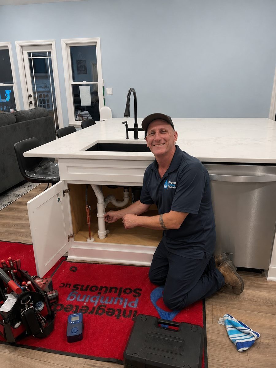 Male technician kneels under a kitchen island sink, tightening white PVC pipes in an open cabinet while tools rest on a red mat nearby.