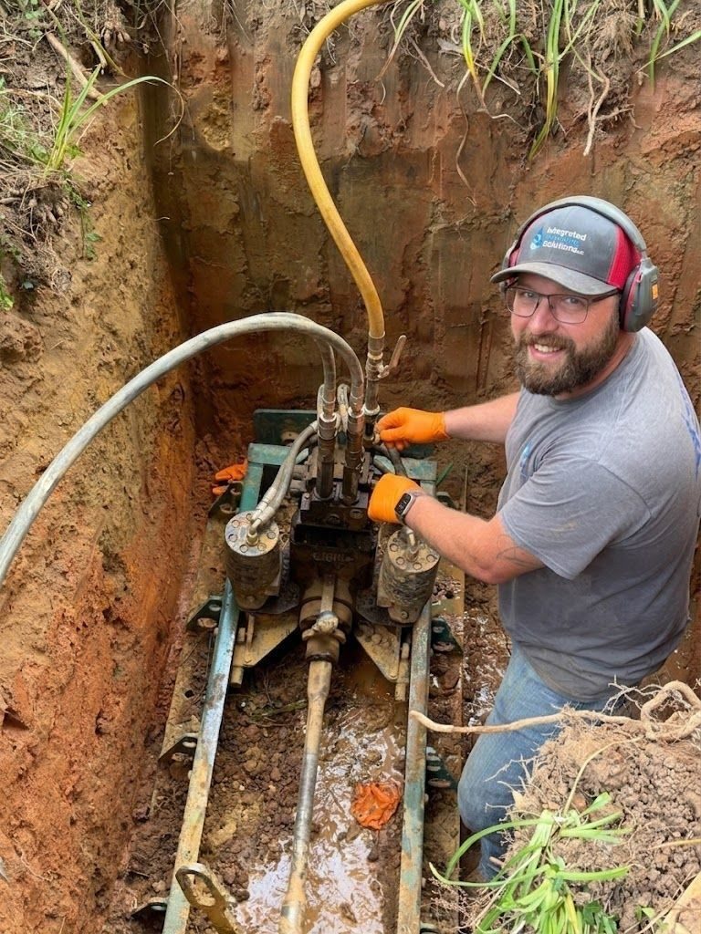 Technician in gloves and ear protection adjusts a hydraulic pump setup in a muddy trench with hoses and metal machinery around him.